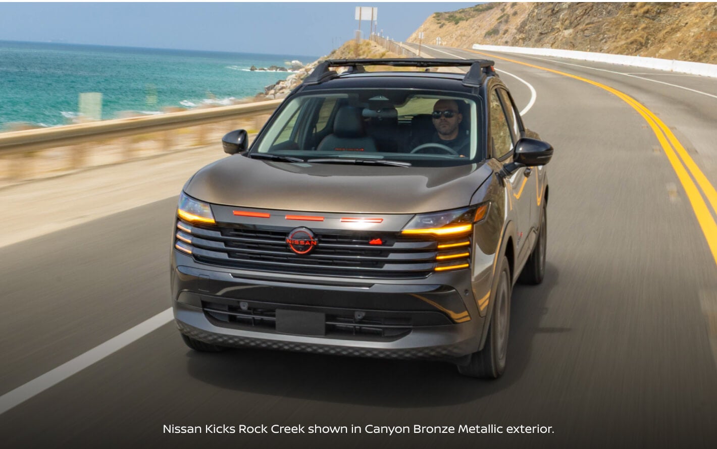 Front view of a 2027 Nissan Kicks Rock Creek in Canyon Bronze Metallic crossing a highway.